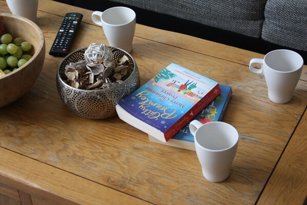 A wooden table with a bowl of green grapes, a basket of dried flowers, a book titled 'The Christmas Project', a remote control, and two white mugs.