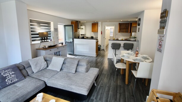 A modern living room with a grey sectional sofa, a dining table with chairs, and a kitchen area in the background. - Bournemouth hot tub and sauna holiday let (Bournemouth)