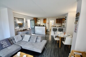 A modern living room with a grey sectional sofa, a dining table with chairs, and a kitchen area in the background.