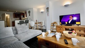 A modern living room with a grey sofa, wooden coffee table, and a television displaying a man in a suit. - Bournemouth hot tub and sauna holiday let (Bournemouth)