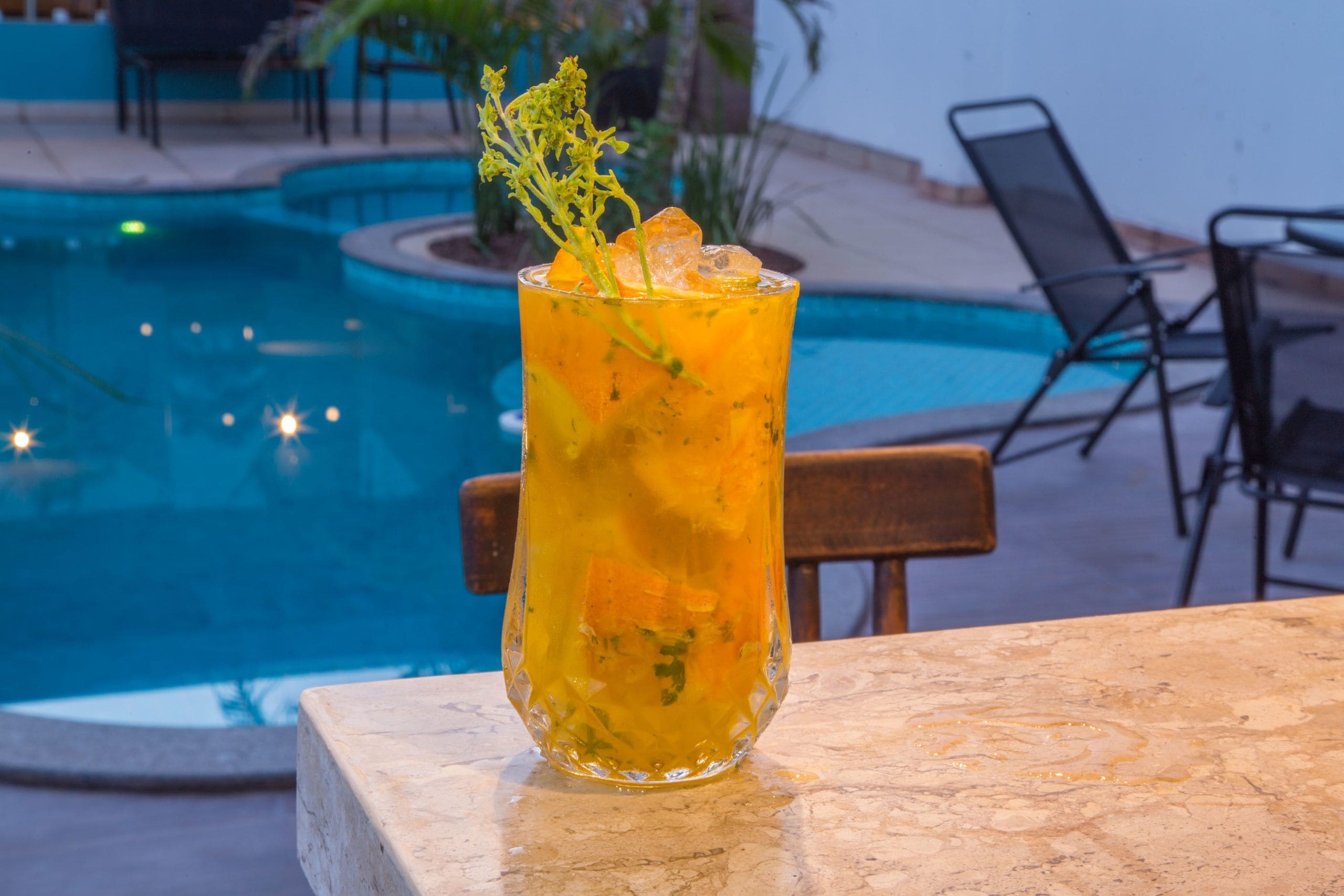 A glass of iced beverage with a garnish on a marble table by a poolside.