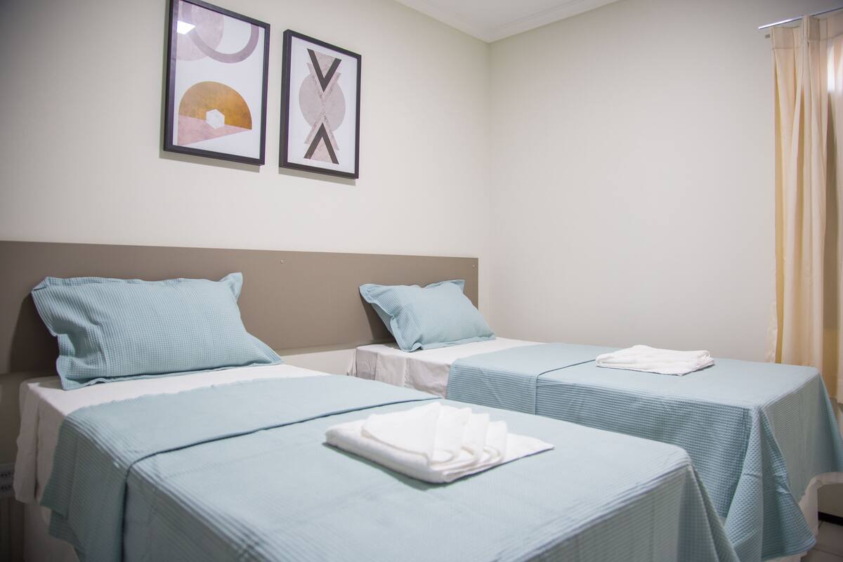 Habitación de hotel con dos camas, cada una con cubrecama y almohadas azules, y una toalla blanca en la cama al frente.