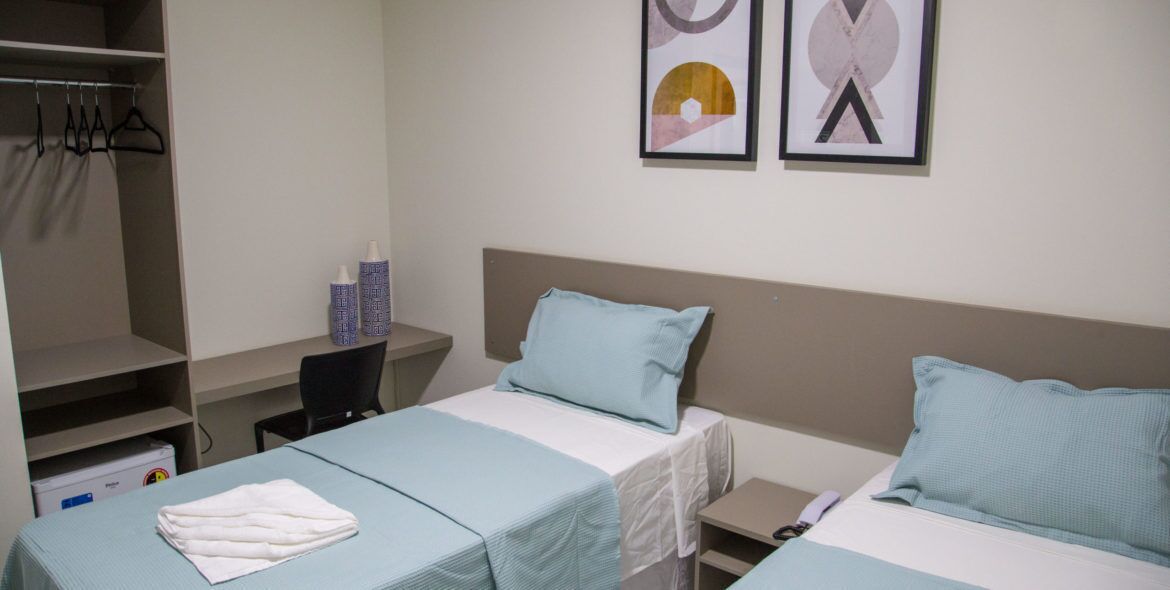 A hotel room with two single beds, a desk, and a chair. There is a wardrobe with hangers and a small table with a lamp.
