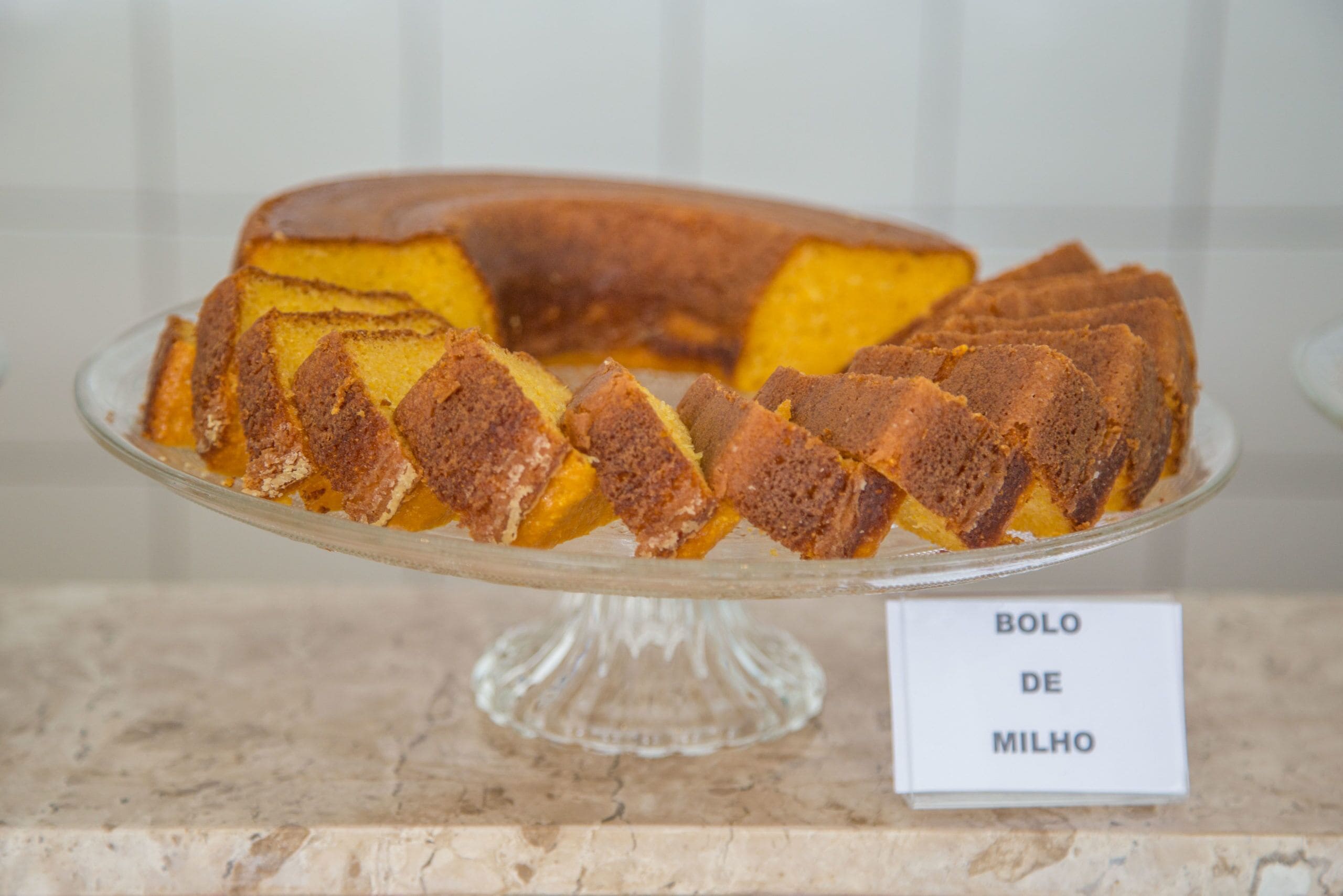 A bundt cake with a label reading "BOLO DE MILHO" on a clear glass stand.
