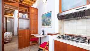 A compact kitchen with a gas stove, wooden cabinets, a small dining table with red chairs, and a painting of boats on the wall.