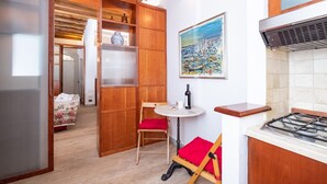 A compact kitchen with wooden cabinets, a small dining table with red cushions, a painting of a harbor, and a gas stove.