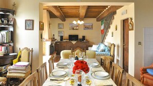 Un comedor con una mesa larga dispuesta para seis personas, sillas de madera, una araña y una cocina al fondo.