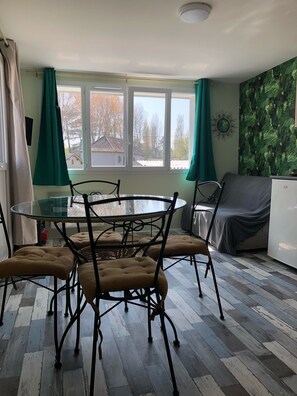 A dining area with a glass-top table, four cushioned chairs, a white refrigerator, and a window with curtains.