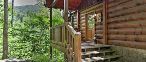 A wooden cabin with a porch and steps leading to a door, surrounded by lush greenery.