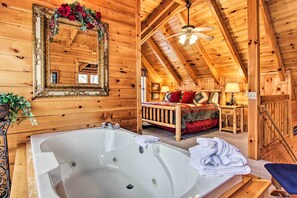 Innenansicht einer Holzhütte mit einer großen Badewanne, einem Bett mit roter Bettwäsche und einem Spiegel mit floralem Dekor.
