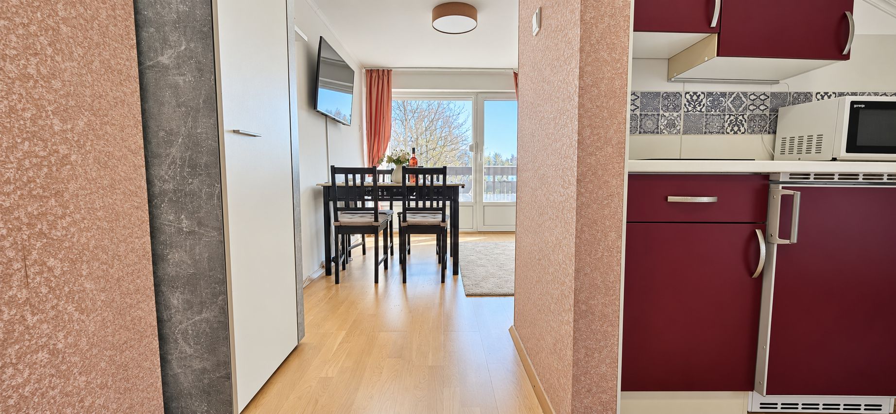 Ein Flur, der zu einem Essbereich mit Tisch und Stühlen sowie zu einer Küche mit roten Schränken und einer Mikrowelle führt, mit Blick ins Freie.