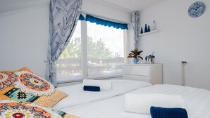 A bedroom with a large bed, a window with curtains, a white dresser, and a small shelf with decorative items.