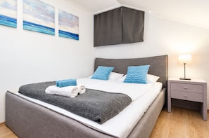 Ein modernes Schlafzimmer mit einem großen Bett, grauer Bettwäsche, blauen Kissen und einem Nachttisch mit einer Lampe.