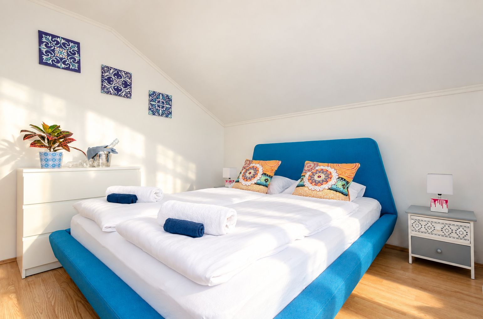 A neatly made bed with white and blue bedding, two decorative pillows, a bedside table with a lamp, and a wall-mounted plant.