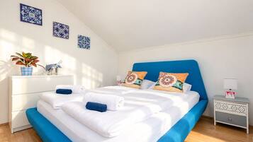 Cama bem arrumada com roupa de cama branca e azul, duas almofadas decorativas, mesa de cabeceira com abajur e planta fixada na parede.