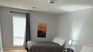 Ein Zimmer mit zwei Betten, einem Fenster mit Jalousien, einem Gemälde und einem Nachttisch mit Lampe.