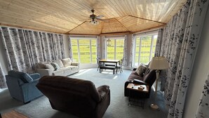 Ein geräumiges Wohnzimmer mit Holzboden, großen Fenstern, einer Couch, einem blauen Sessel und einem Tisch mit Lampe.