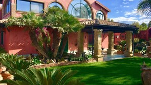 A pink house with large arched windows, a covered patio, and a well-maintained garden with palm trees. - Holiday home Villa Flumini  (Quartu Sant'Elena)