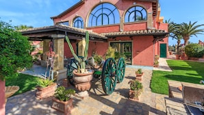 A two-story house with a red facade, large arched windows, and a courtyard with potted plants and a green lawn. - Holiday home Villa Flumini  (Quartu Sant'Elena)