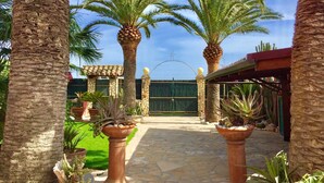 A courtyard with palm trees, a stone pathway, and a building with a green gate. - Holiday home Villa Flumini  (Quartu Sant'Elena)