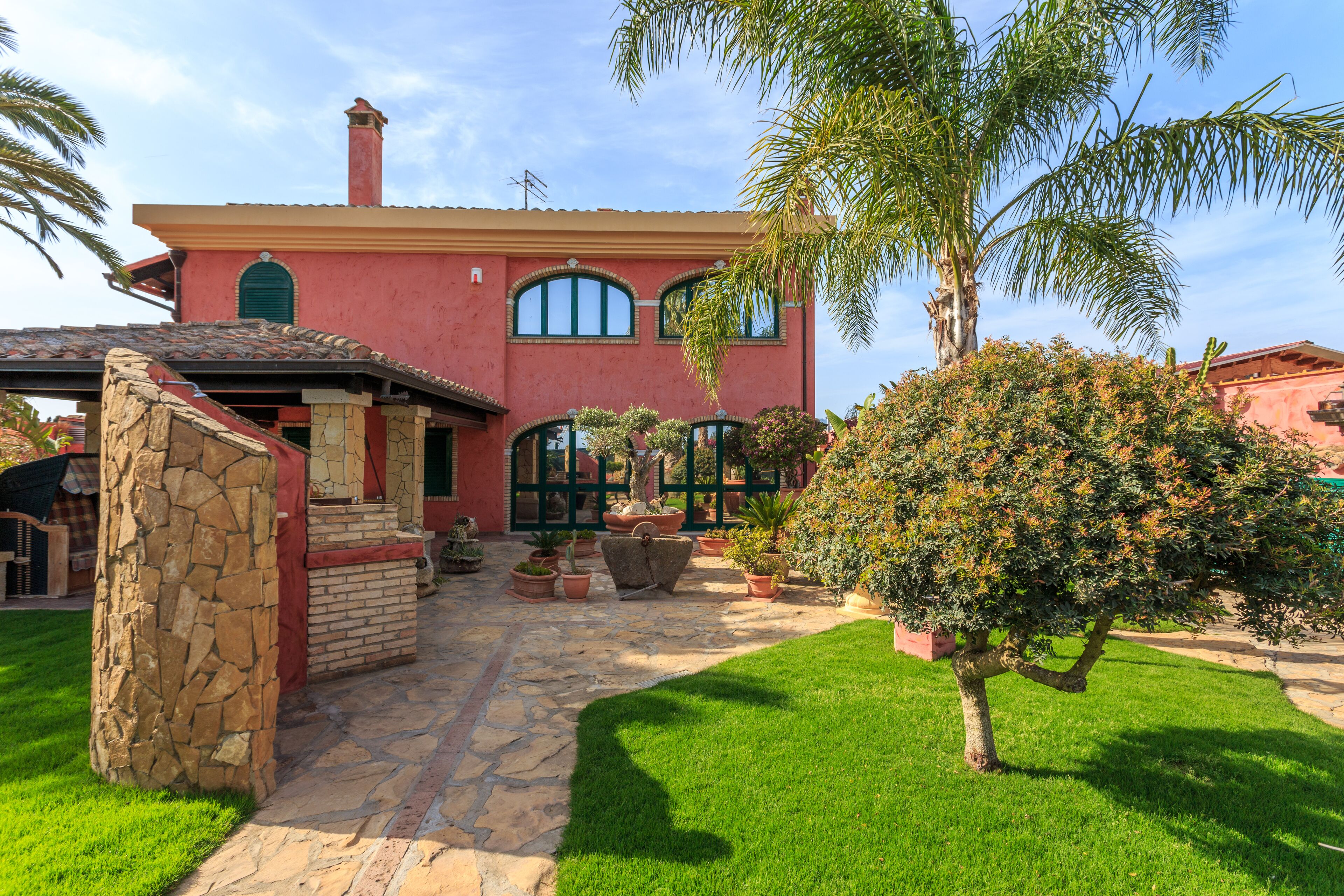 A house with a stone facade, a tiled roof, and a well-maintained garden with various plants and a palm tree.
