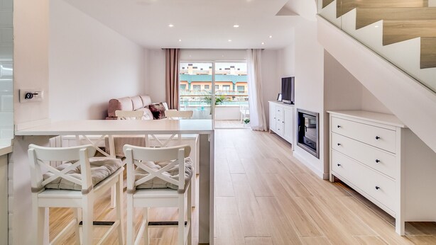 Una cocina moderna con armarios blancos, un comedor con sillas blancas, una escalera de madera y vistas a edificios a través de amplios ventanales.