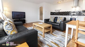 Un soggiorno moderno con un divano in pelle nera, un tavolino in legno e una TV. È presente una zona cucina con schienale nero e bianco e un piano cottura bianco.