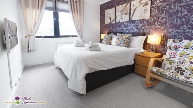 Un dormitorio con cama, mesita de noche, lámpara y un cuadro floral en la pared.