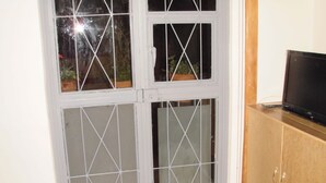 Une pièce dotée d’une porte-fenêtre coulissante donnant sur un balcon, d’un meuble en bois sur lequel est posée une télévision, et d’un revêtement de sol en tissu à motifs.