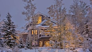 A large, multi-story lodge with a prominent balcony and a lit interior, surrounded by snow-covered trees.