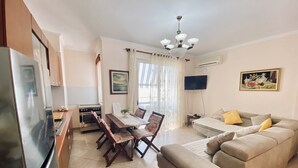 A living room with a sofa, a dining table with chairs, a kitchen area with a refrigerator, oven, and a TV mounted on the wall. - Fantastic Beach View at ARIEL’s HOUSE (Durrës)