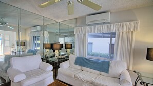 Un salon avec un canapé blanc, une table basse et une table à manger en verre, ainsi que des chaises. On y trouve également un ventilateur de plafond, un appareil de climatisation et une fenêtre avec des rideaux.