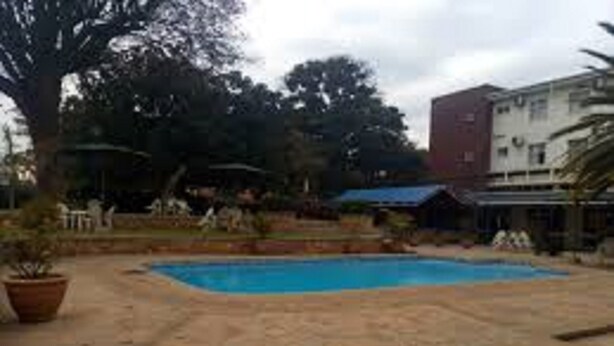 A swimming pool with a stone deck, surrounded by trees and a building in the background. - Royal Cashrise lodge (Kabwe)