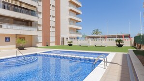 A swimming pool with a tiled edge, surrounded by a building with balconies and a clear sky. - PIROPO - Apartment with sea views in Playa de Gandia. (Grau i Platja)