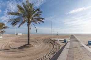 A sandy beach with a palm tree, a paved walkway, and a distant view of the sea. - PIROPO - Apartment with sea views in Playa de Gandia. (Grau i Platja)