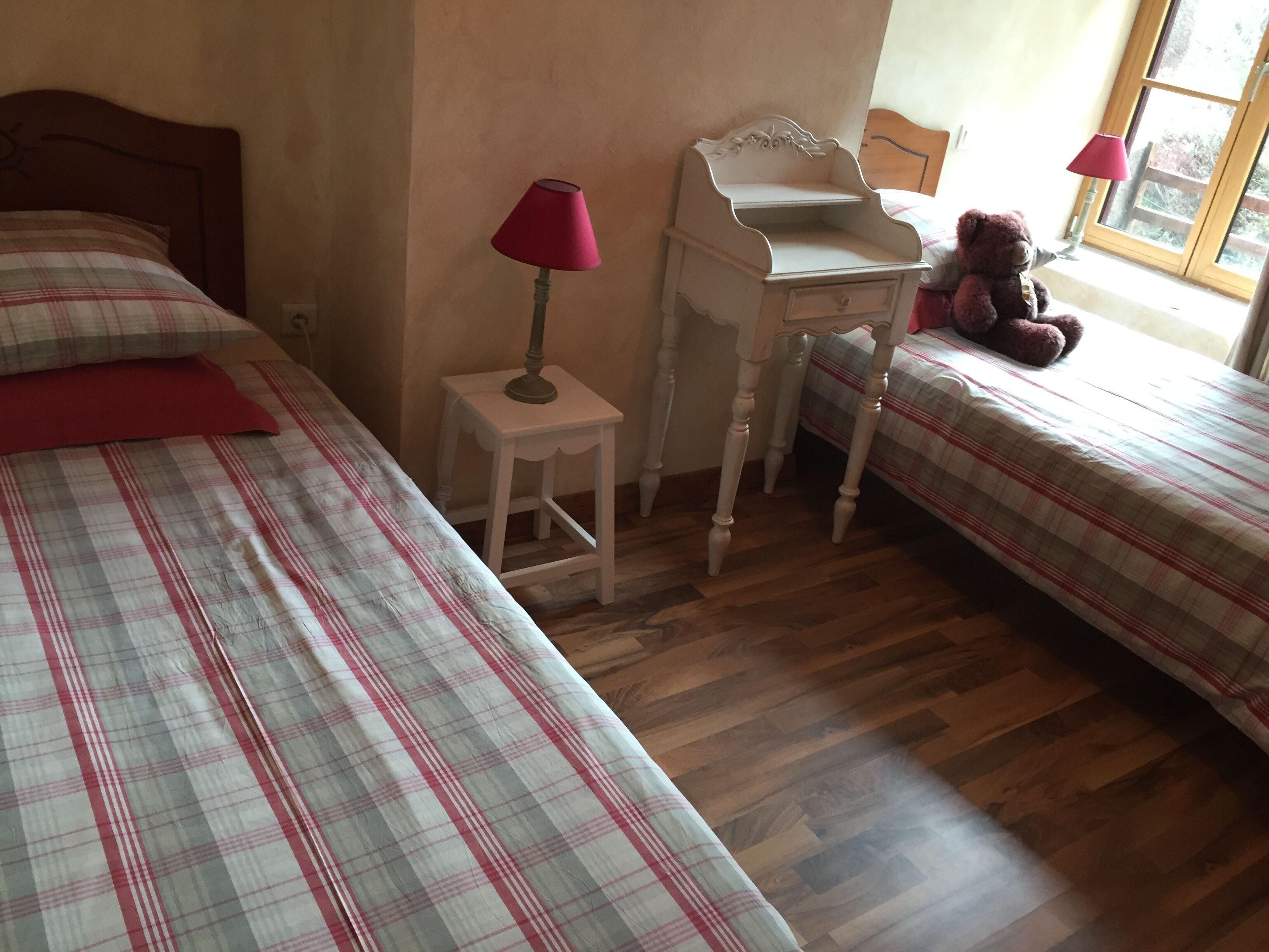 Une chambre avec deux lits, un petit bureau, une chaise, une lampe et un ours en peluche.