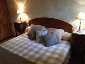 Cama bem arrumada com colcha xadrez e várias almofadas, com duas mesas de cabeceira com abajures, em quarto com cabeceira de madeira e parede lisa.