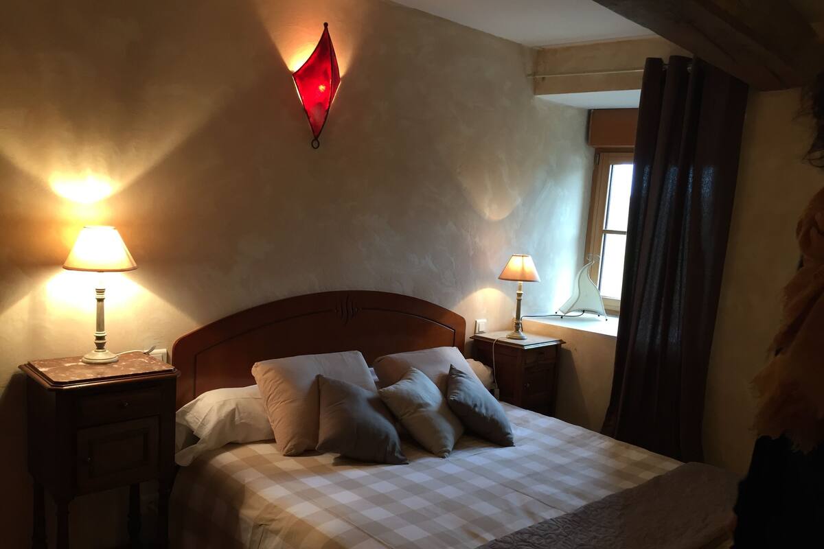 A neatly made bed with a checkered bedspread and multiple pillows, flanked by two bedside tables with lamps, one of which has a red light fixture on the wall.