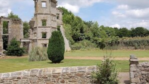 Un edificio histórico de piedra con una torre, rodeado por un césped cuidado y muros de piedra.