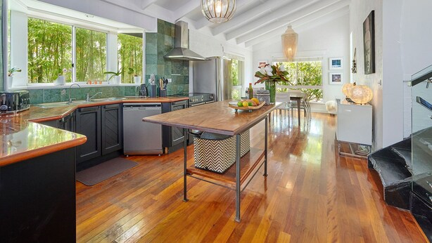 A modern kitchen with a central island, wooden flooring, and large windows offering a view of greenery.