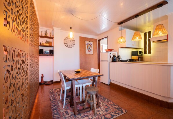 A cozy kitchen with a wooden table, white chairs, and a patterned rug. There is a decorative wall panel and a clock on the wall.