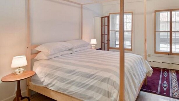 A four-poster bed with a wooden frame and white bedding. There is a small bedside table with a lamp and a decorative rug on the floor. - Pizo Lodge (Kabwe)