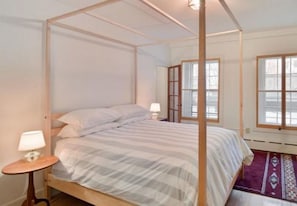 Una cama con dosel de madera y ropa de cama blanca. Hay una mesita de noche pequeña con una lámpara y una alfombra decorativa en el suelo.
