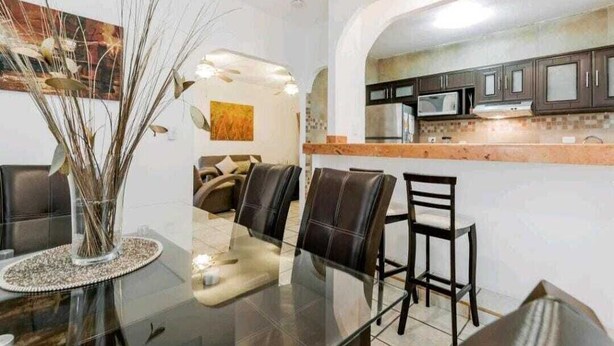 A modern kitchen with dark cabinets and a microwave, a dining area with a glass table and leather chairs, and a decorative vase with branches.