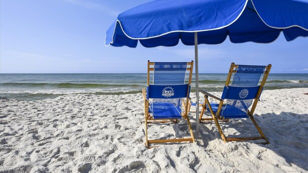 Deux chaises de plage sous un parasol bleu, sur une plage de sable, avec l’océan en arrière-plan.