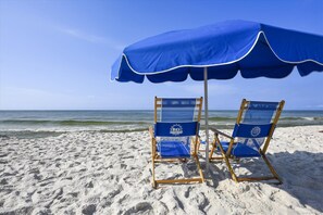 Two beach chairs under a blue umbrella on a sandy beach with the ocean in the background.