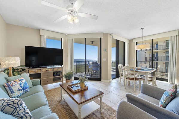A spacious living room with a flat-screen TV, comfortable seating, a dining area, and a balcony with a view.