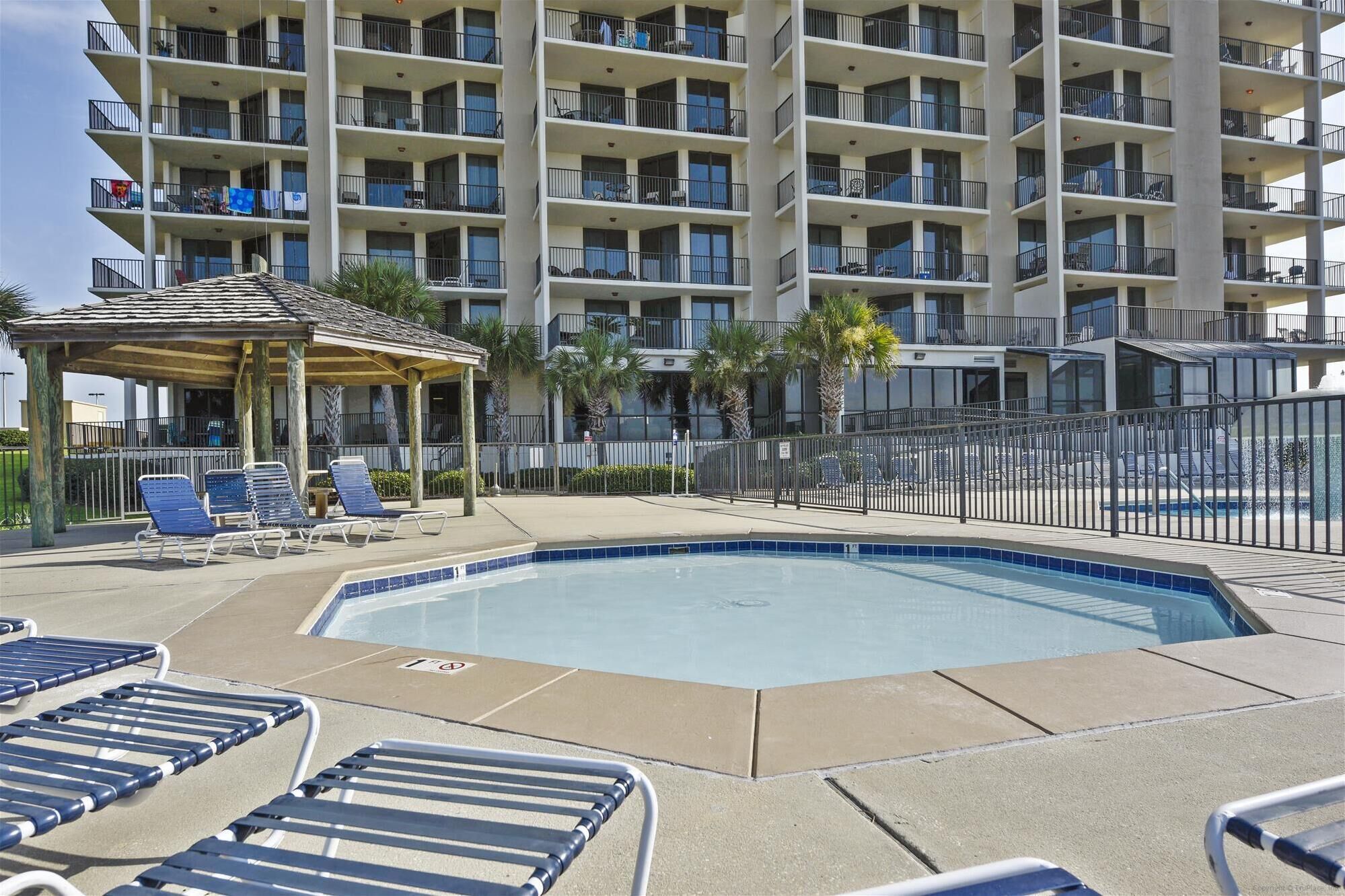 A swimming pool with lounge chairs, a covered seating area, and a multi-story building with balconies in the background.