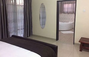 Une chambre à coucher avec un lit, un miroir, une fenêtre avec des rideaux et une salle de bain visible à travers une porte.