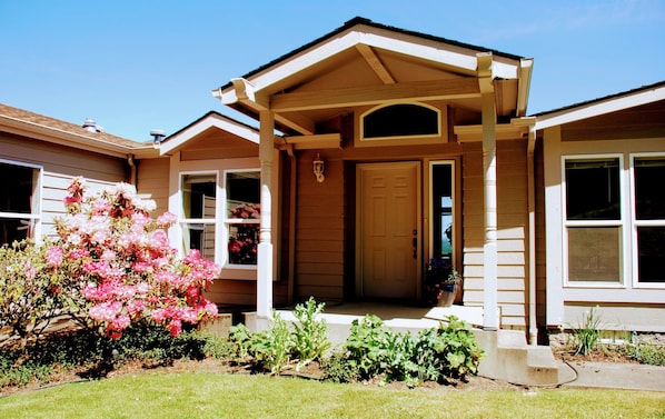 A two-story house with a covered porch, a front door, and a small garden with pink flowers. - Sarver Winery Guesthouse (Eugene)